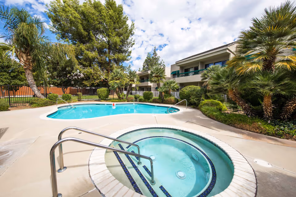 Outdoor area of Citrus Place featuring a swimming pool and a hot tub surrounded by palm trees and other greenery, with a two-story building in the background under a partly cloudy sky.