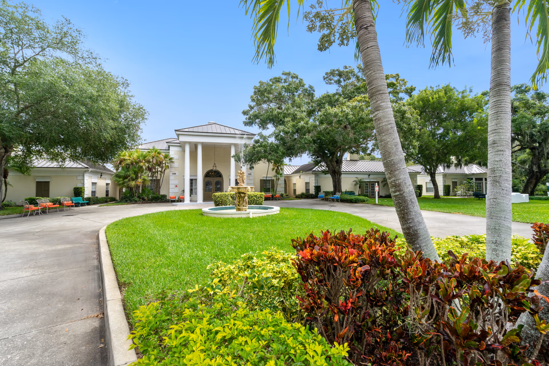 Front entrance of a senior living building with a circular driveway, central fountain, and landscaped lawns and palm trees.