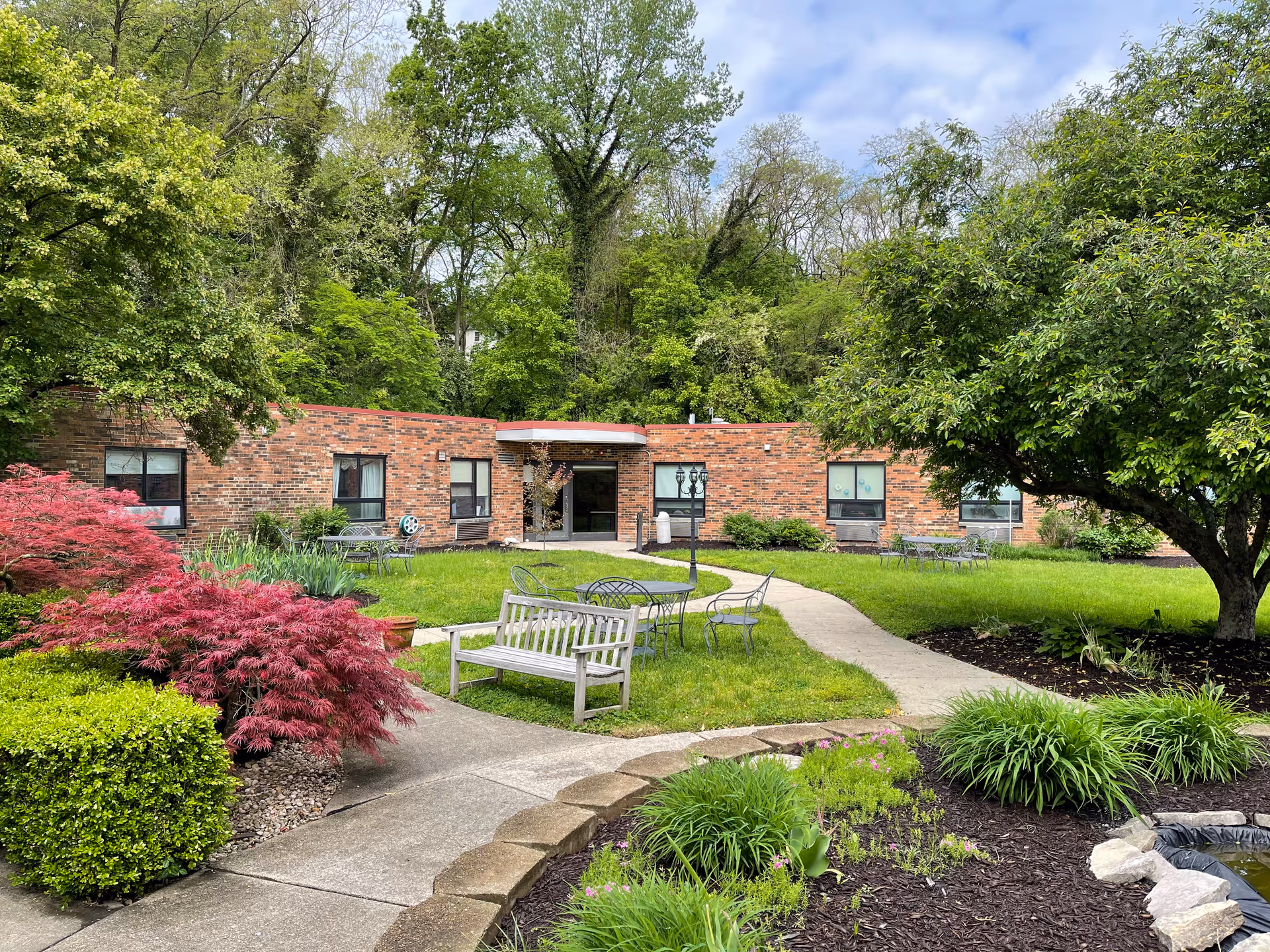 Courtyard of a single-story brick senior living building with benches, metal chairs and tables, a walking path, and landscaped trees and shrubs.