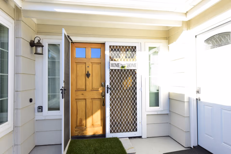 Entrance area with a wooden front door partially open behind a white security screen door that has a sign reading 'Home Sweet Home'. The area is well-lit with natural light, has beige siding walls, a small patch of artificial grass on the floor, and a wall-mounted lantern light fixture on the left side.