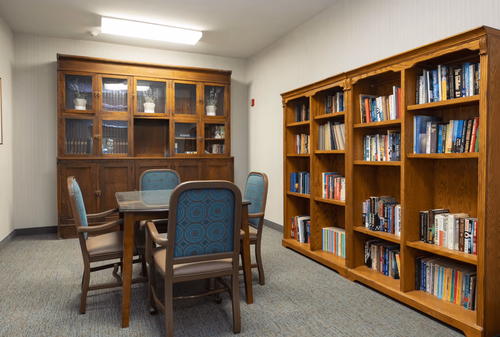 A small room with a wooden table surrounded by four chairs with blue patterned upholstery. On the right side, there are wooden bookshelves filled with books. On the back wall, there is a wooden cabinet with glass doors containing some potted plants and books. The room has beige walls and carpeted flooring.