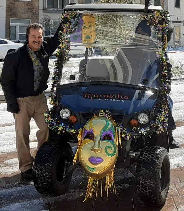 A man stands next to a decorated golf cart with a Mardi Gras-style mask attached to the front. The golf cart has the text 'Maravilla @ The Domain' on its front. The scene is outdoors with snow on the ground and buildings in the background.