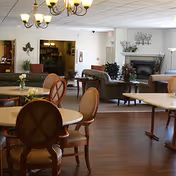 A spacious common area in an assisted living facility featuring round tables with chairs, a chandelier, and a seating area with sofas and armchairs near a fireplace. The room has wooden flooring and is decorated with plants and wall art.
