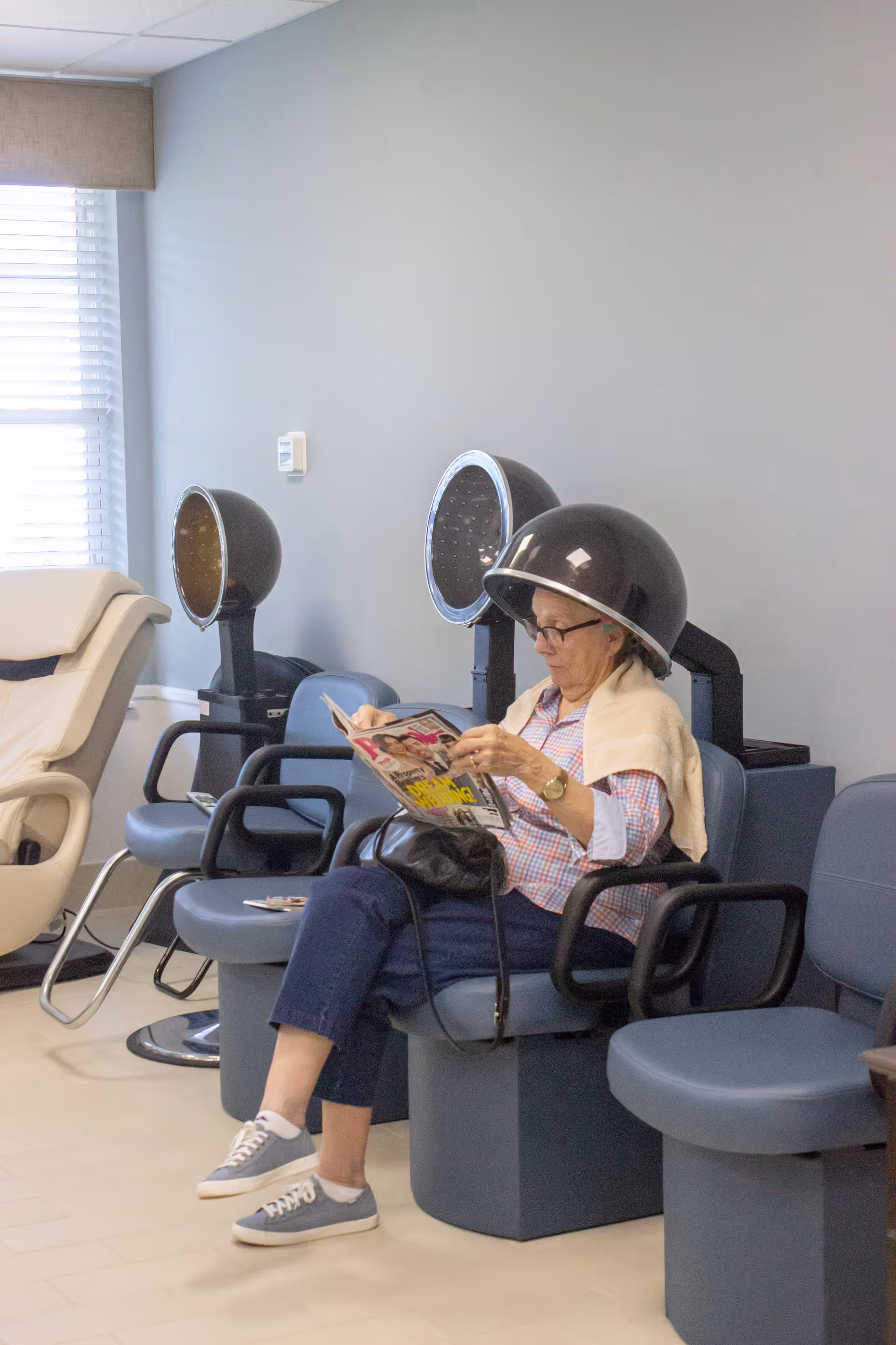 An elderly woman sitting under a hair dryer in a salon area, reading a magazine. She is wearing glasses, a checkered shirt, and blue pants, with a towel draped over her shoulders. The room has several salon chairs and hair dryers, with a window letting in natural light.