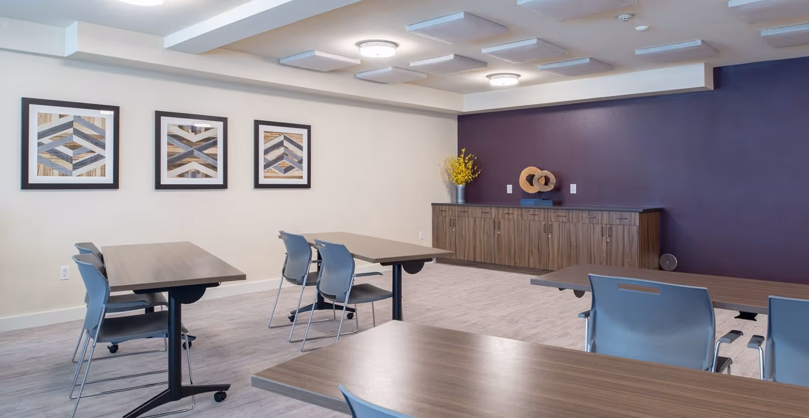 A modern dining room with several wooden tables and gray chairs arranged neatly. The room features a purple accent wall with a wooden cabinet decorated with a vase of yellow flowers and a circular sculpture. Three framed geometric art pieces hang on a white wall.