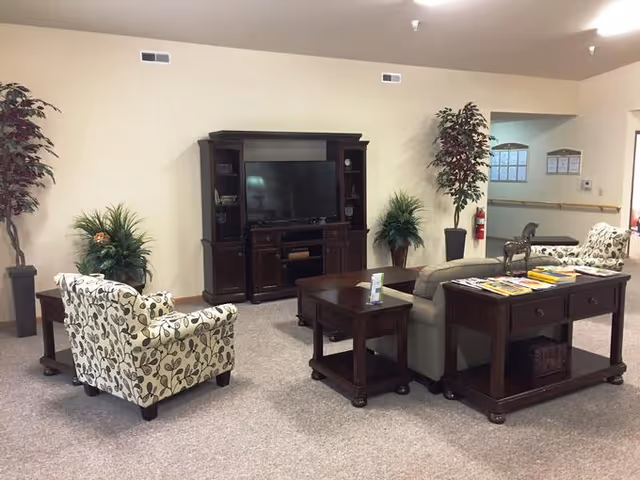 A cozy living room area in an assisted living facility featuring a patterned armchair, a beige sofa, dark wooden tables, and a dark wooden entertainment center with a flat-screen TV. There are potted plants placed around the room, and a fire extinguisher is mounted on the wall near an open doorway.