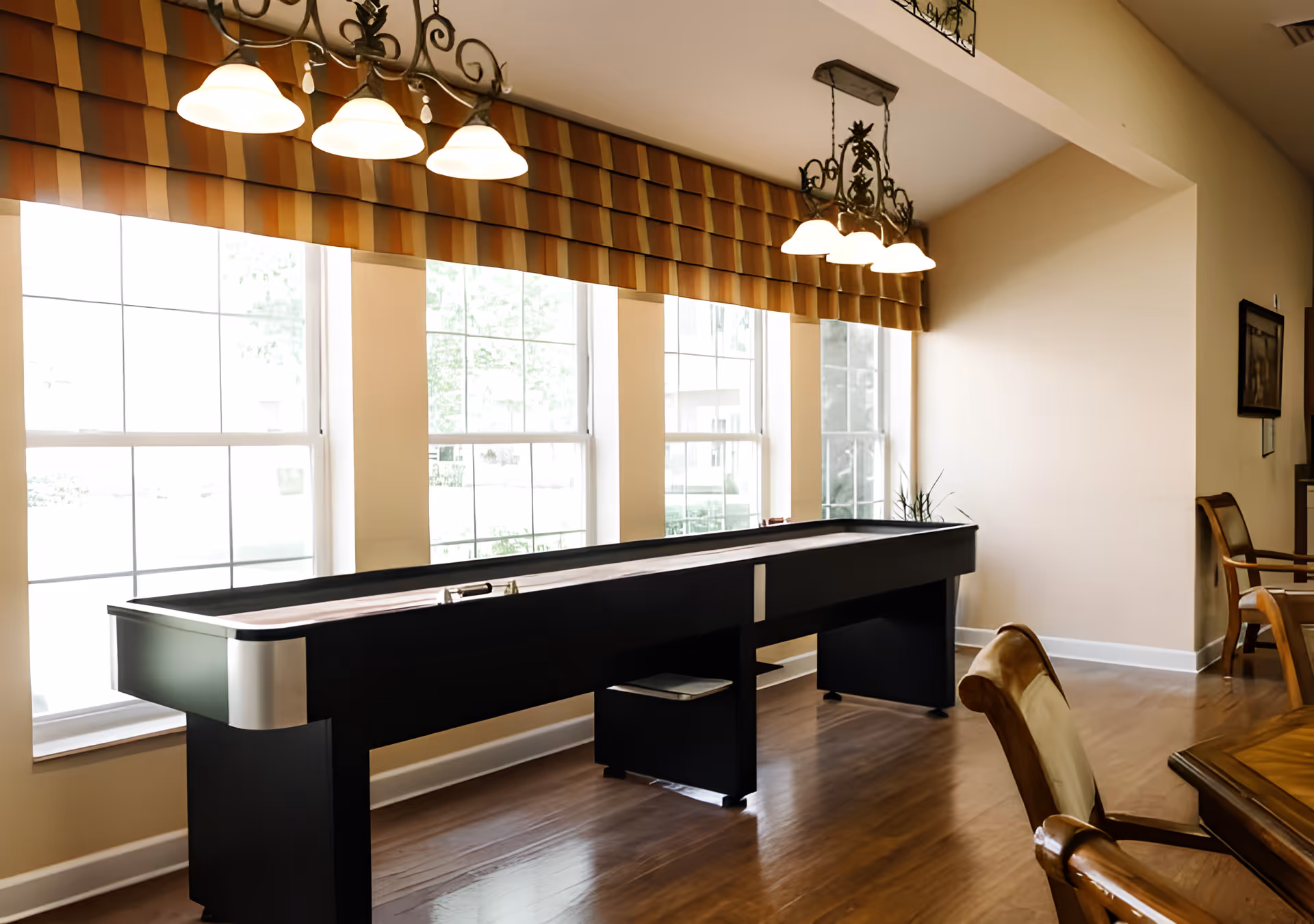 A well-lit room with large windows covered by striped valances, featuring a long black shuffleboard table in the center. The room has wooden flooring, a wooden table with cushioned chairs, and decorative hanging light fixtures above the shuffleboard table.