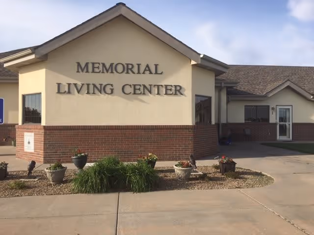 Exterior view of the Memorial Living Center building showing a beige and brick facade with the facility name prominently displayed on the front wall. There are several potted plants and landscaping in front of the building, and a clear sky above.