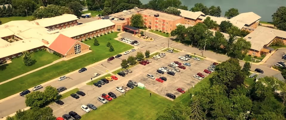 Aerial view of Bethany on the Lake senior living facility showing multiple connected buildings with a large parking lot filled with cars, surrounded by green lawns and trees, and a body of water in the background.