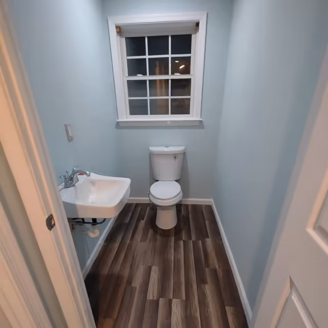 Small bathroom with light blue walls, a white toilet centered under a window with white trim, and a white wall-mounted sink on the left side. The floor has wood-patterned vinyl or laminate flooring.