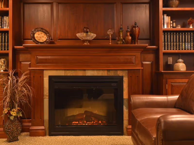 A cozy living room area featuring a wooden fireplace mantel with decorative items on top, built-in bookshelves filled with books and ornaments on either side, a leather armchair to the right, and a decorative vase with dried plants on the left.