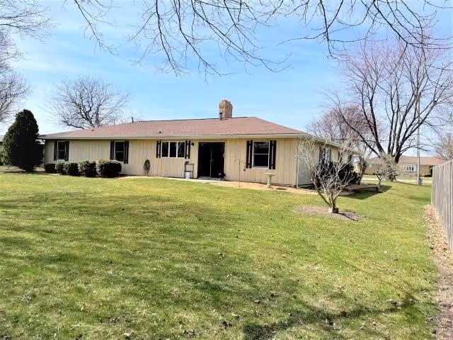 Single-story building with beige siding and a brown roof, surrounded by a large grassy lawn with some bushes and leafless trees under a clear blue sky.