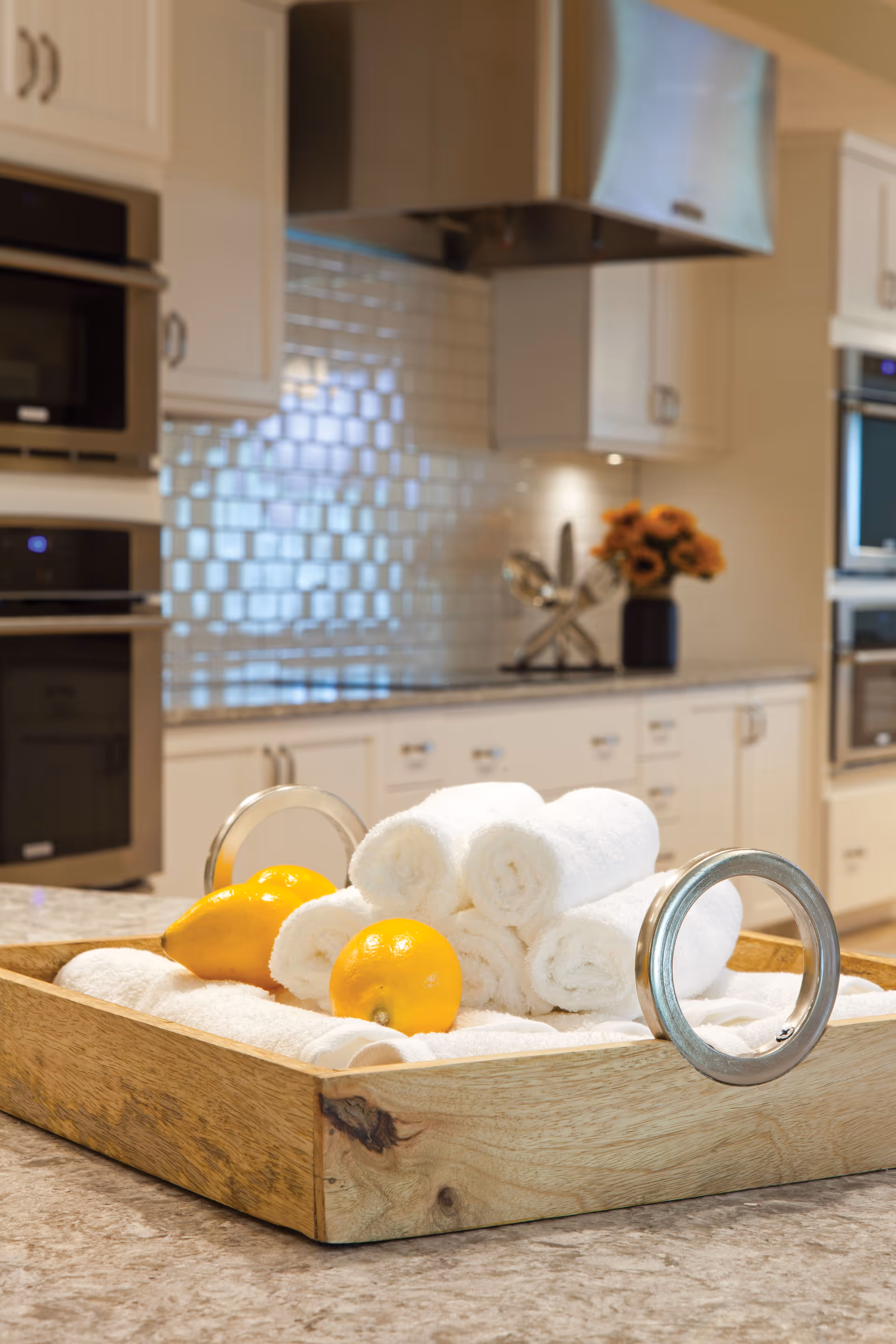 A wooden tray on a kitchen countertop holding rolled white towels and two yellow lemons. The background shows a modern kitchen with white cabinets, stainless steel appliances, and a tiled backsplash.