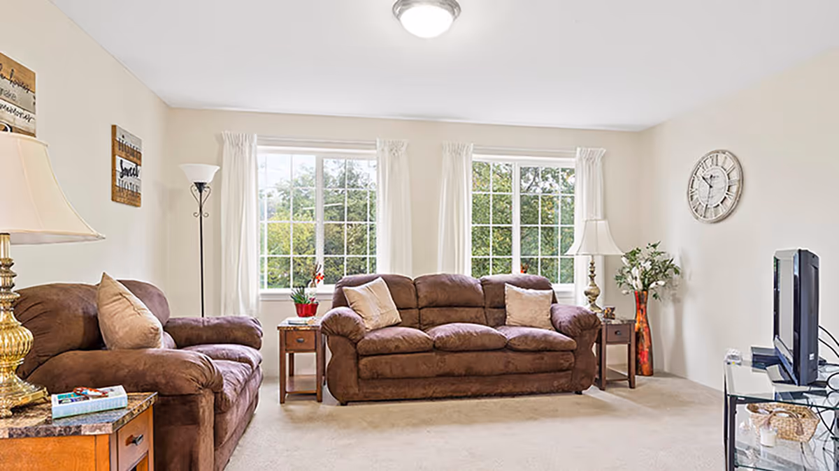 Sunlit living room with a brown upholstered sofa and armchair, side tables and lamps, a TV, and large windows overlooking greenery.
