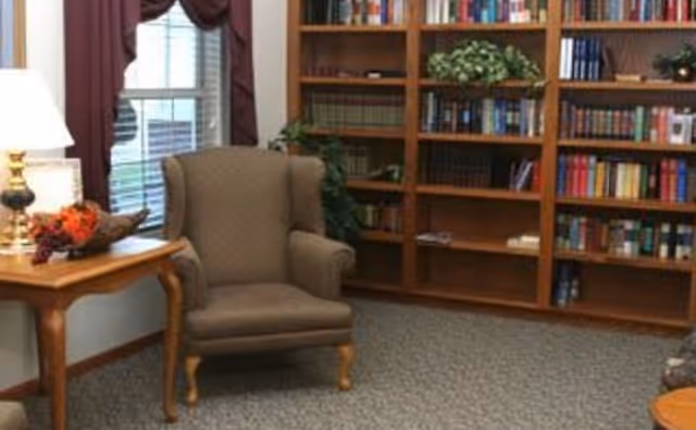 Cozy reading area with an upholstered armchair beside a lamp-lit side table and wall-length bookshelves.