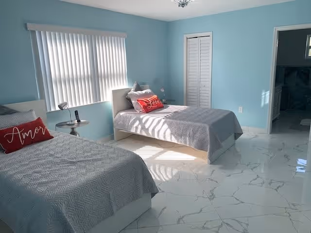 A bright bedroom with two single beds, each with gray bedding and a red decorative pillow with white text. The walls are painted light blue, and there is a window with vertical blinds letting in sunlight. A small round side table with a lamp is positioned between the beds. The floor is covered with white marble tiles, and there is a white closet door and an open doorway leading to another room.