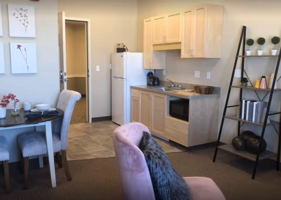 Senior living apartment interior with dining table, pink chair, kitchenette, and shelving unit.