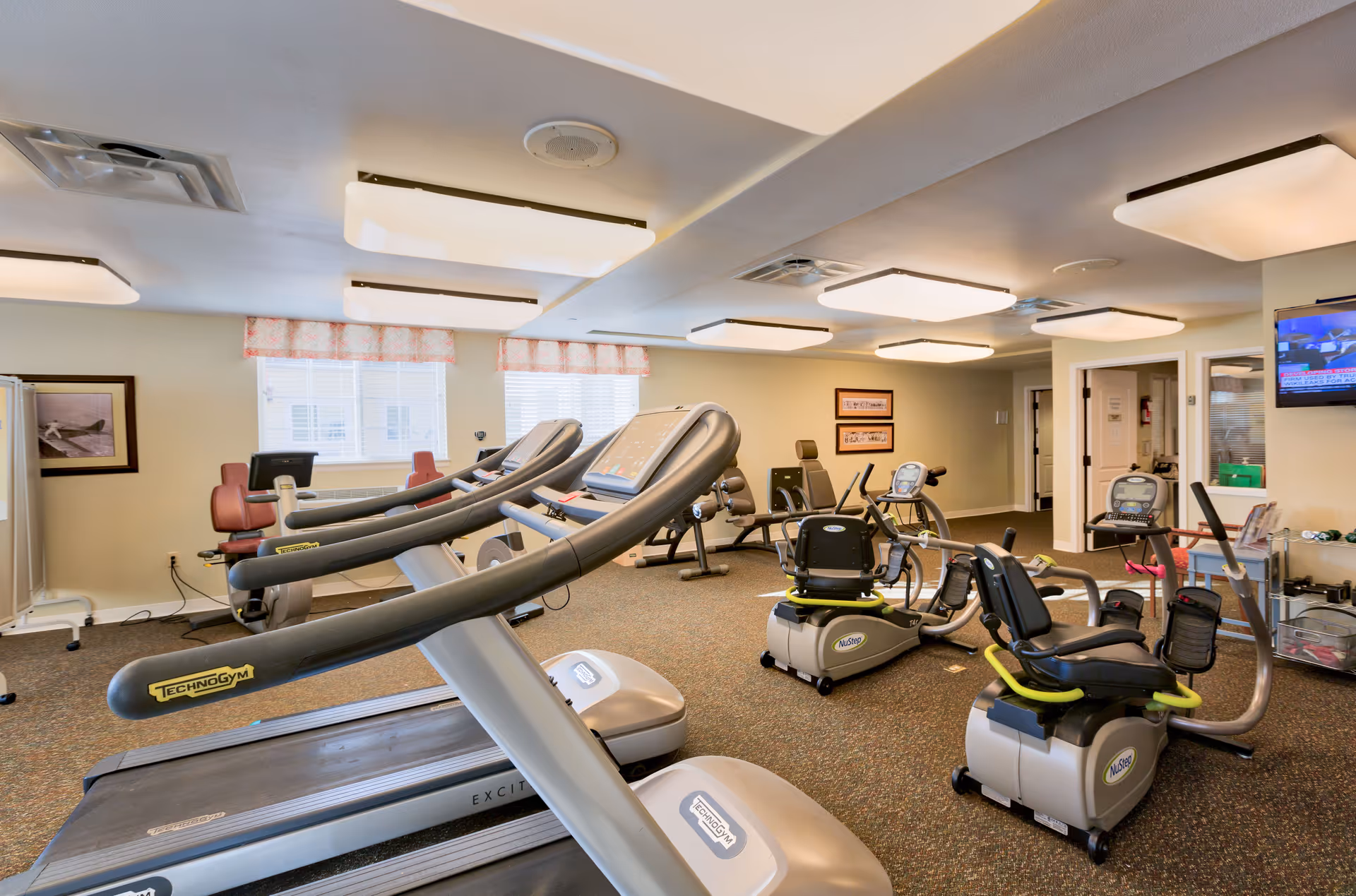 Interior view of a senior living facility fitness room with multiple exercise machines including treadmills and recumbent bikes. The room is well-lit with ceiling lights and has windows with patterned valances. There is a television mounted on the wall and framed pictures on the walls.