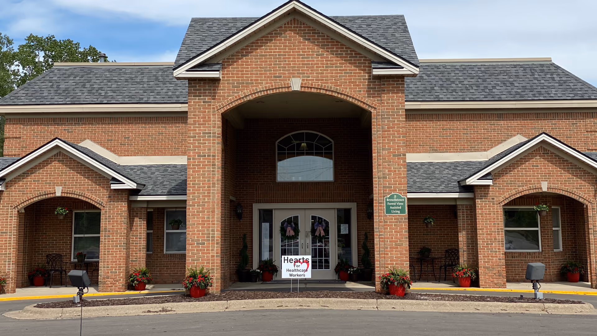 Brick two-story assisted living building front with a covered entrance, double glass doors, and potted plants.