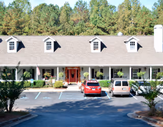 Front exterior of a single-story assisted living community with a covered porch, two parked cars, and trees behind it.