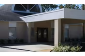 Exterior view of a building entrance with double oval glass doors under a covered driveway. The building has beige walls, small windows on either side of the doors, and some landscaping with bushes and grass in front.
