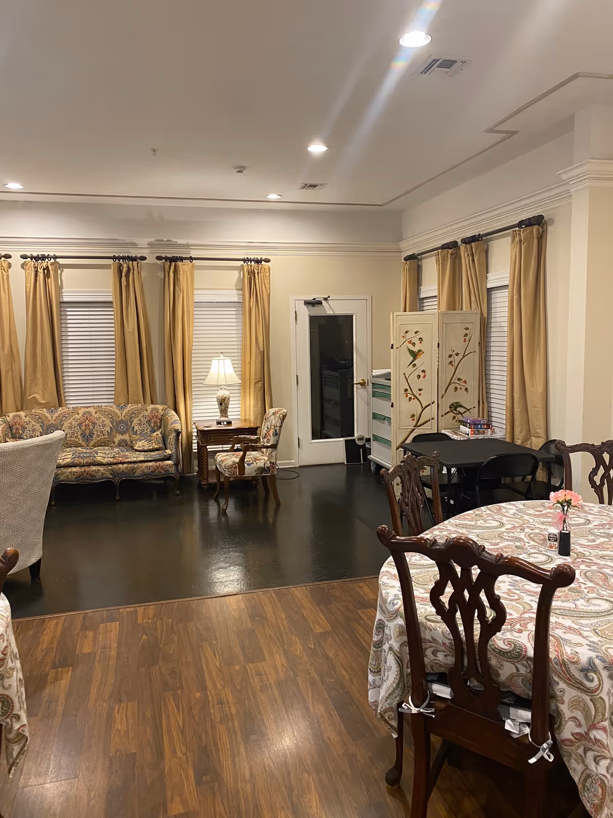Open common room with round dining tables and chairs, a seating area with a floral sofa and lamp, and curtained windows.