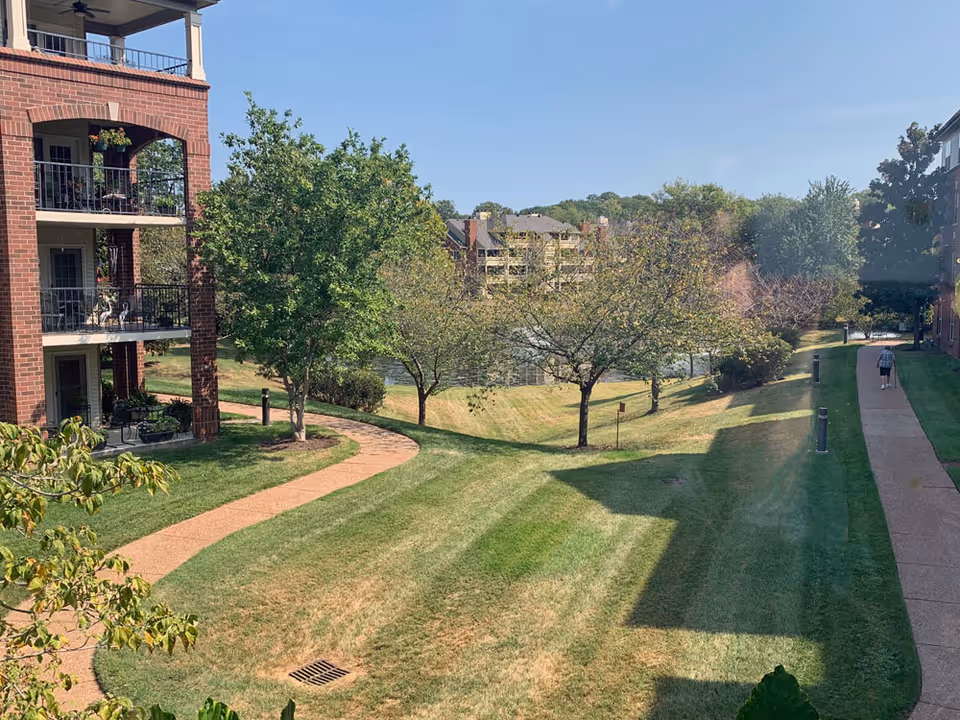View of a landscaped outdoor area at a senior living facility with green grass, trees, a pond in the background, and two brick buildings on either side. A person is walking on a paved path on the right side of the image under a clear blue sky.