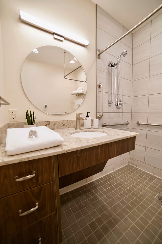 A modern bathroom with a beige tiled walk-in shower equipped with grab bars and a handheld showerhead. There is a wooden vanity with a marble countertop, an undermount sink, a round mirror above the sink, a small plant, and neatly folded white towels on the counter.
