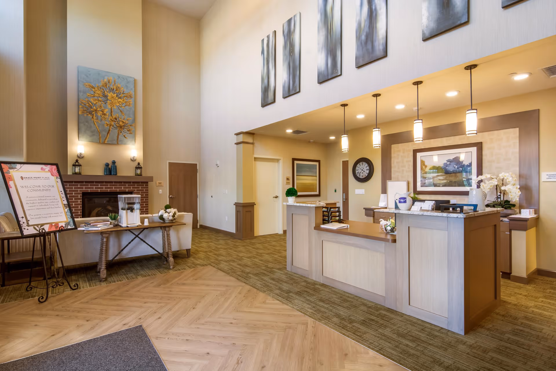 Interior view of a senior living facility reception area with a front desk, pendant lights, wall art, a fireplace with decorative items, a table with water pitchers and cups, and a welcome sign on an easel.