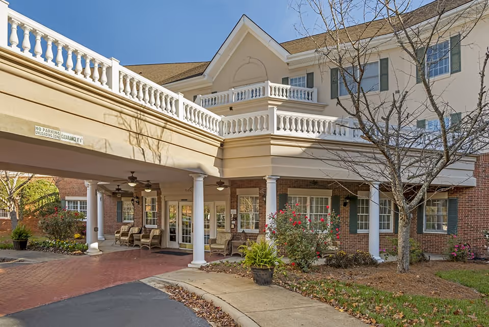 Exterior view of Brookdale South Charlotte senior living facility showing the entrance with a covered drop-off area supported by white columns, outdoor seating with wicker chairs, landscaped garden with bushes and a tree without leaves, and a multi-story building with beige and brick facade under a clear blue sky.