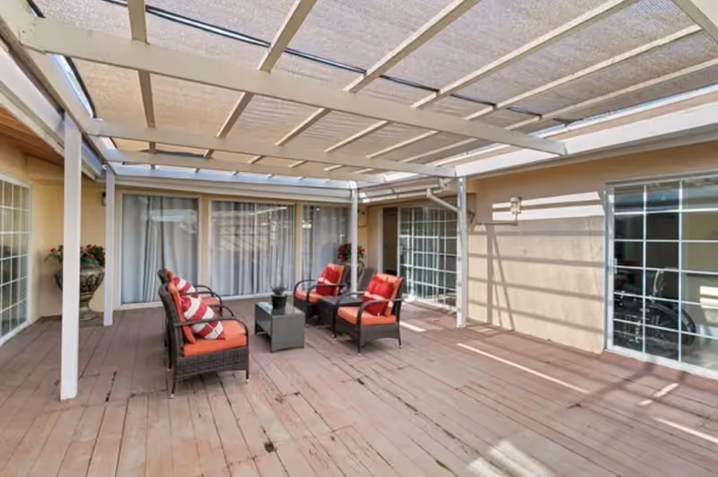 Outdoor patio area with wooden deck flooring, covered by a pergola with a fabric shade. There are four cushioned wicker chairs with red cushions and striped pillows arranged around a small rectangular coffee table. The patio is surrounded by beige walls with large windows and sliding glass doors.