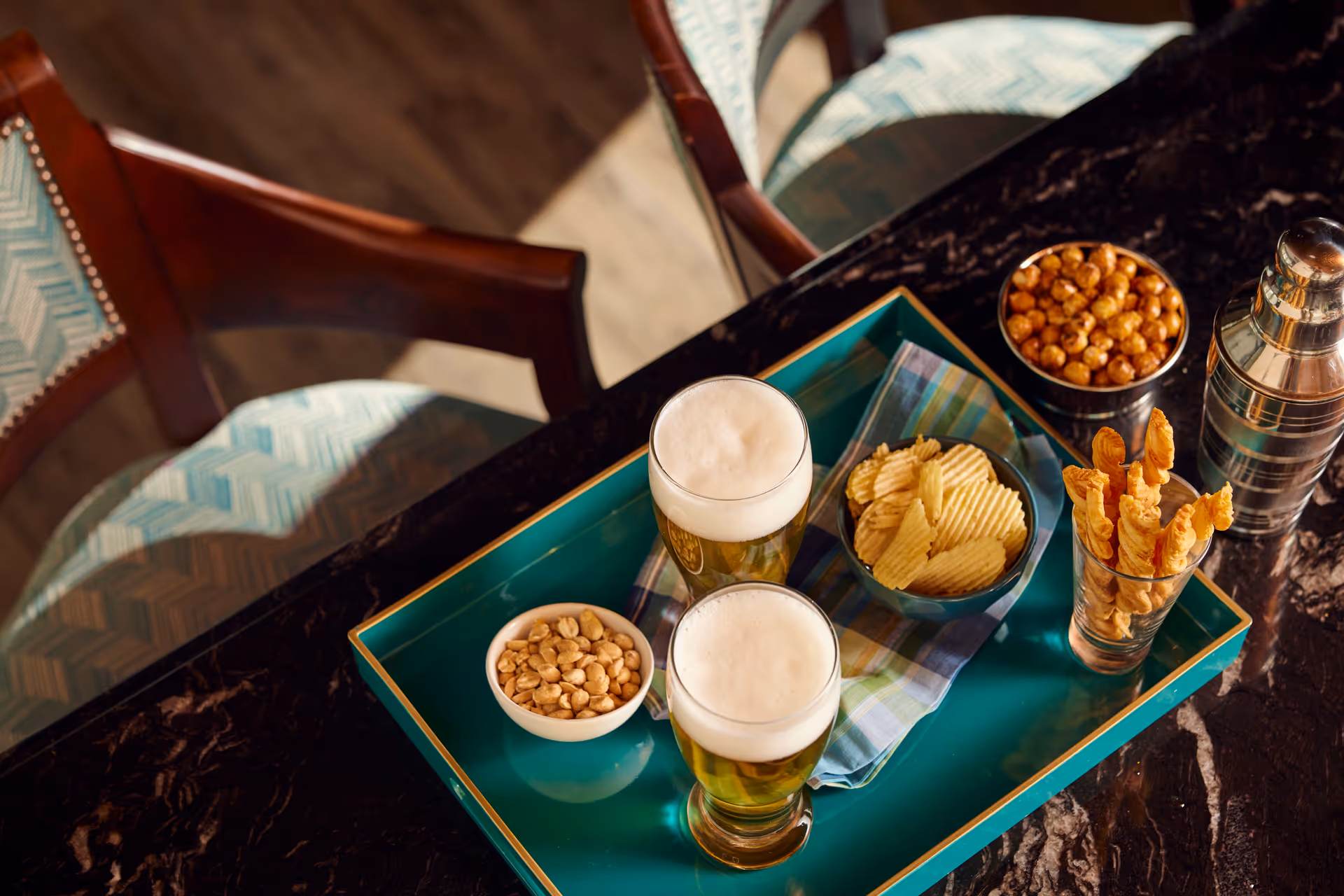 A tray on a dark marble table holding two glasses of beer with foam, a bowl of peanuts, a bowl of potato chips, a glass with breadsticks, and a bowl of roasted chickpeas. Two chairs with patterned cushions are partially visible around the table.