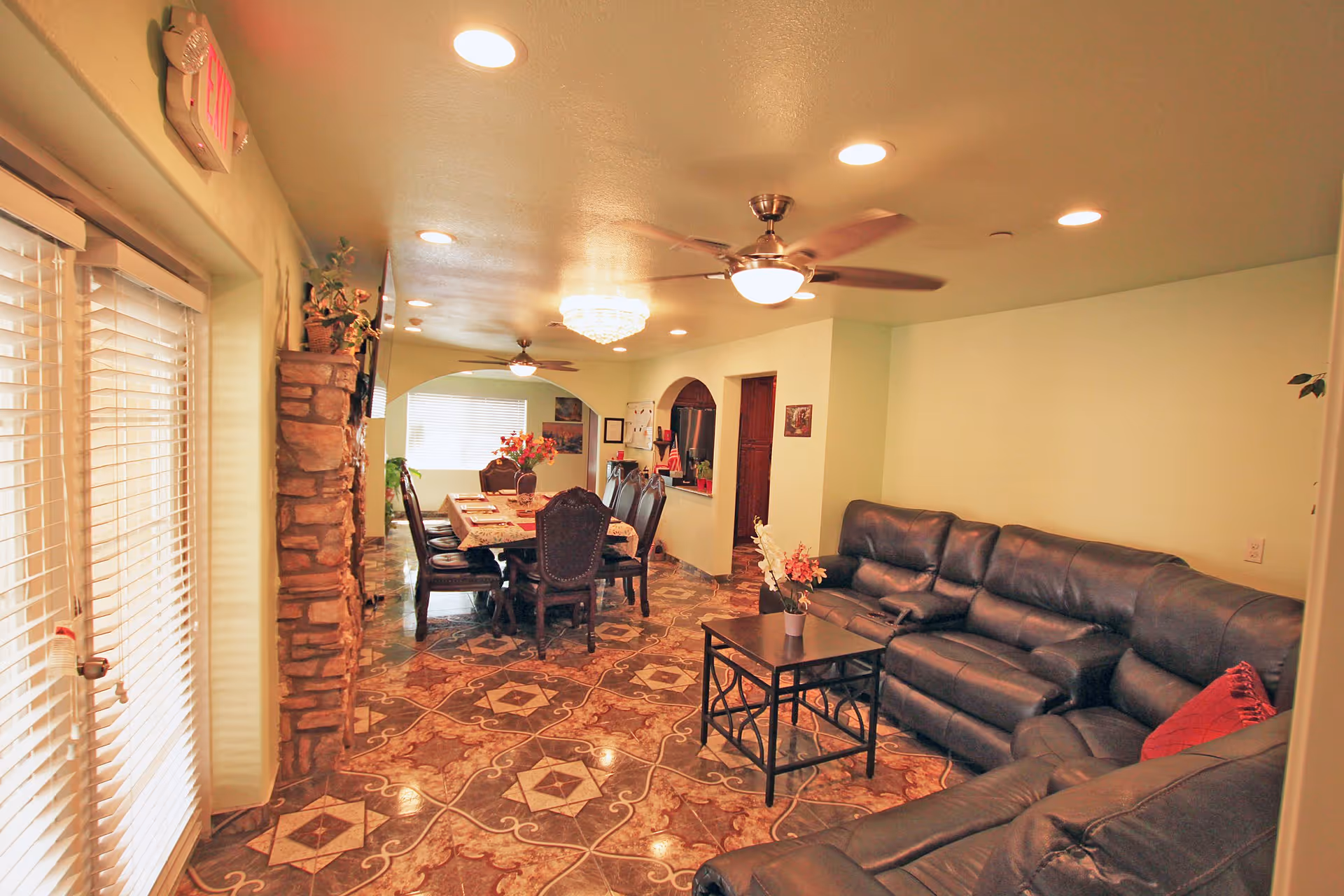 Interior view of a living and dining area in an assisted living facility. The room features a large black leather sectional sofa with a small black coffee table in front, decorated with a small flower arrangement. The dining area has a long table covered with a tablecloth and surrounded by dark wooden chairs. The floor has a patterned tile design, and the ceiling has recessed lighting, ceiling fans, and a chandelier. There is a stone accent wall near a glass door with blinds, and the walls are painted light green.