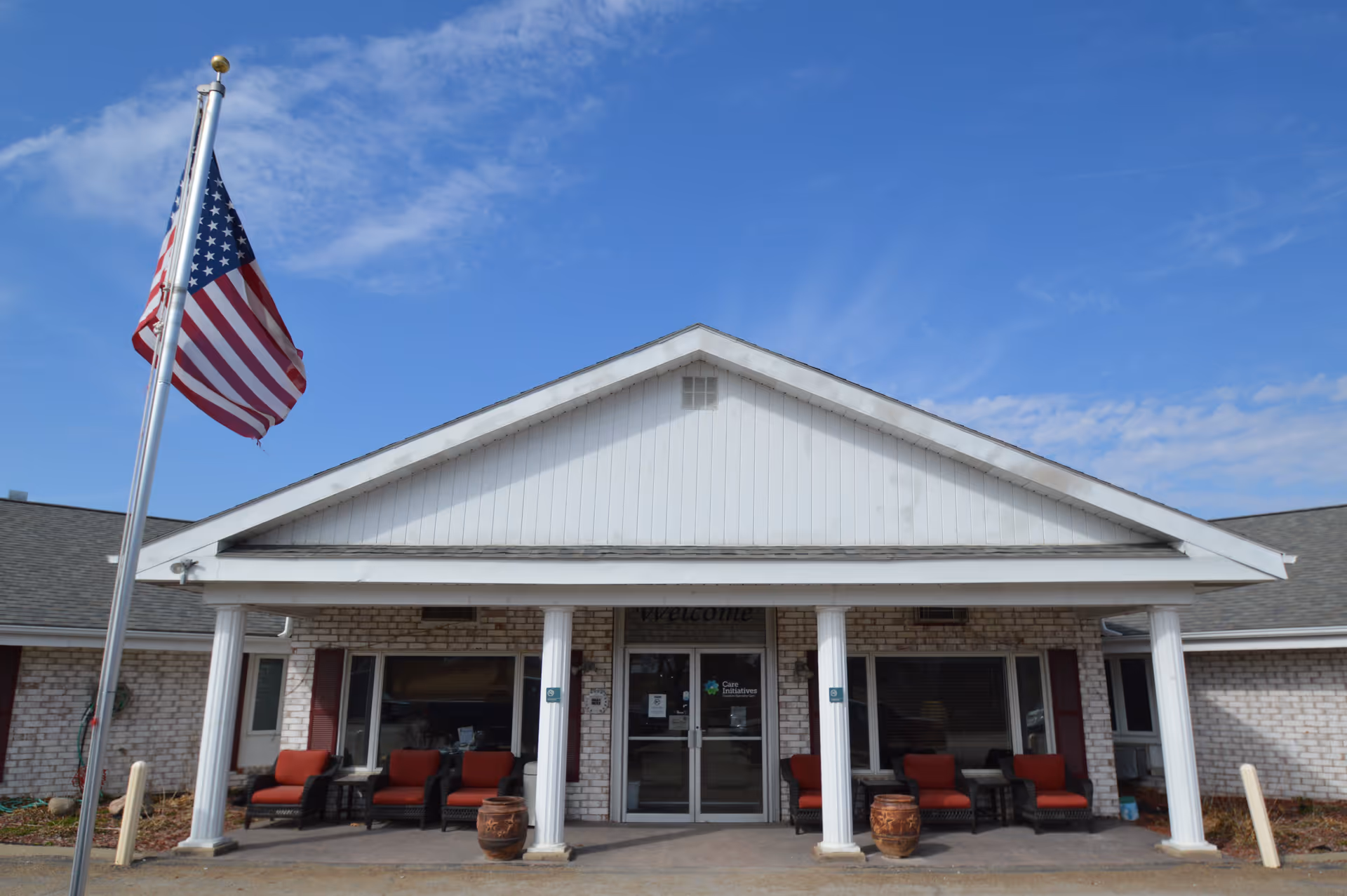 Front exterior view of a single-story brick building with a white triangular roof section supported by four white columns. There are red cushioned chairs and small tables on the porch area. An American flag on a flagpole is visible on the left side against a blue sky with some clouds.