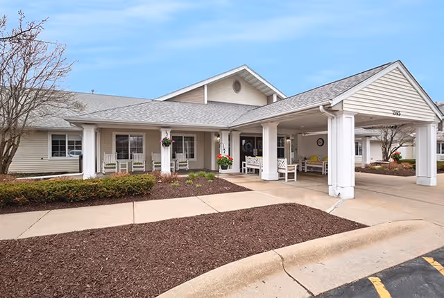 Exterior view of a single-story senior living facility with a covered entrance supported by white columns. The building has beige siding, a gray shingled roof, and several windows. There are benches and hanging flower pots under the covered entrance, with a landscaped area featuring mulch and shrubs in front.