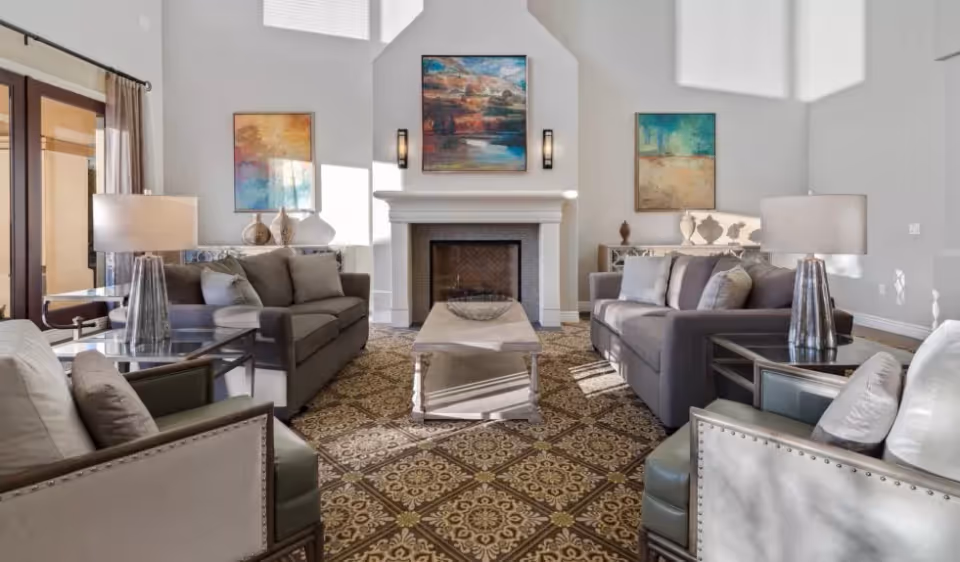 Bright communal living room with sofas and armchairs arranged around a central coffee table and a fireplace, accented with lamps and artwork.