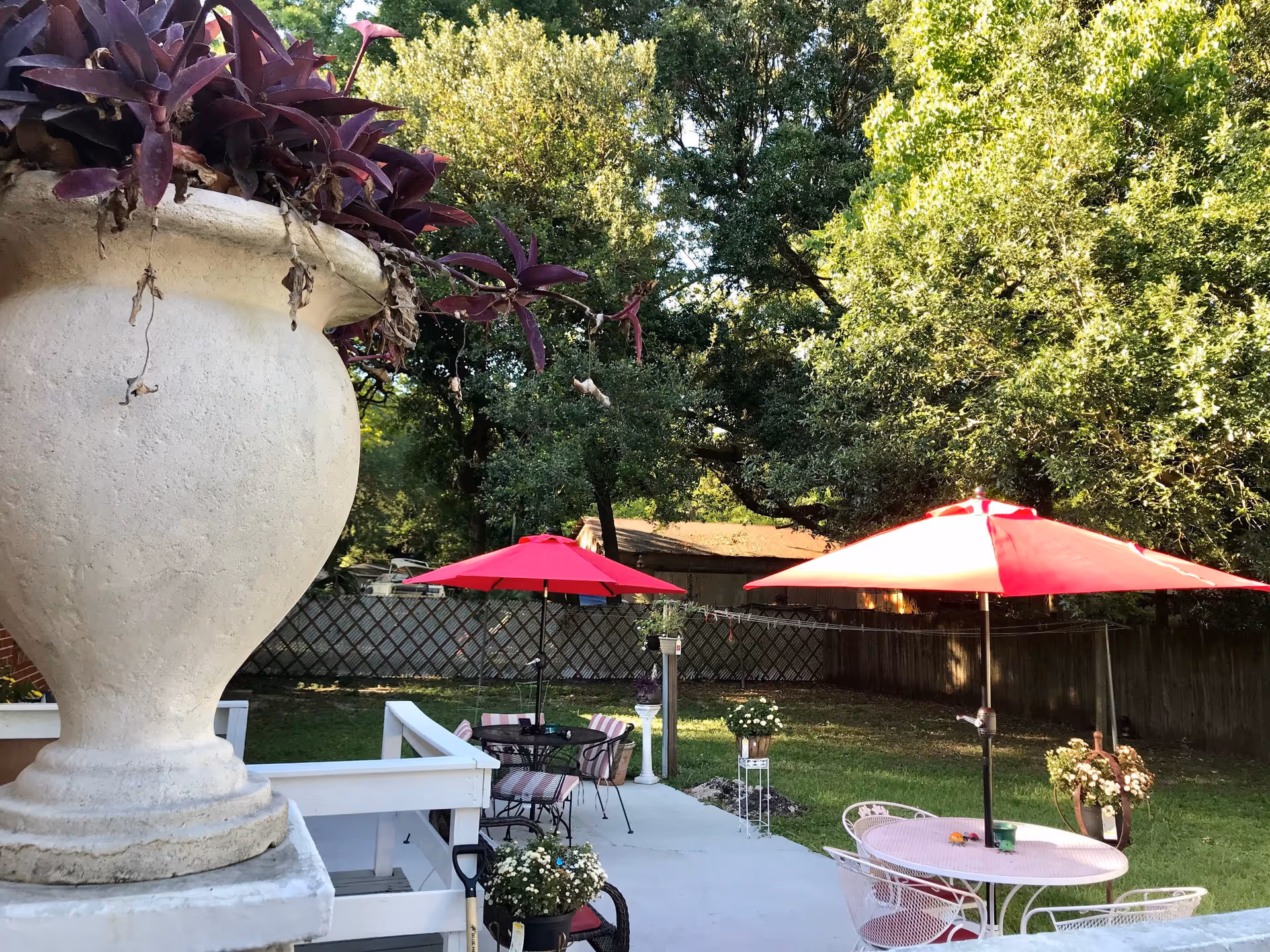 Backyard patio with red umbrellas over outdoor tables and chairs, potted plants, and large trees in the background.
