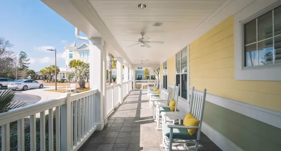 Covered front porch with white railing and rocking chairs with yellow cushions along a yellow-painted building overlooking a driveway and landscaping.