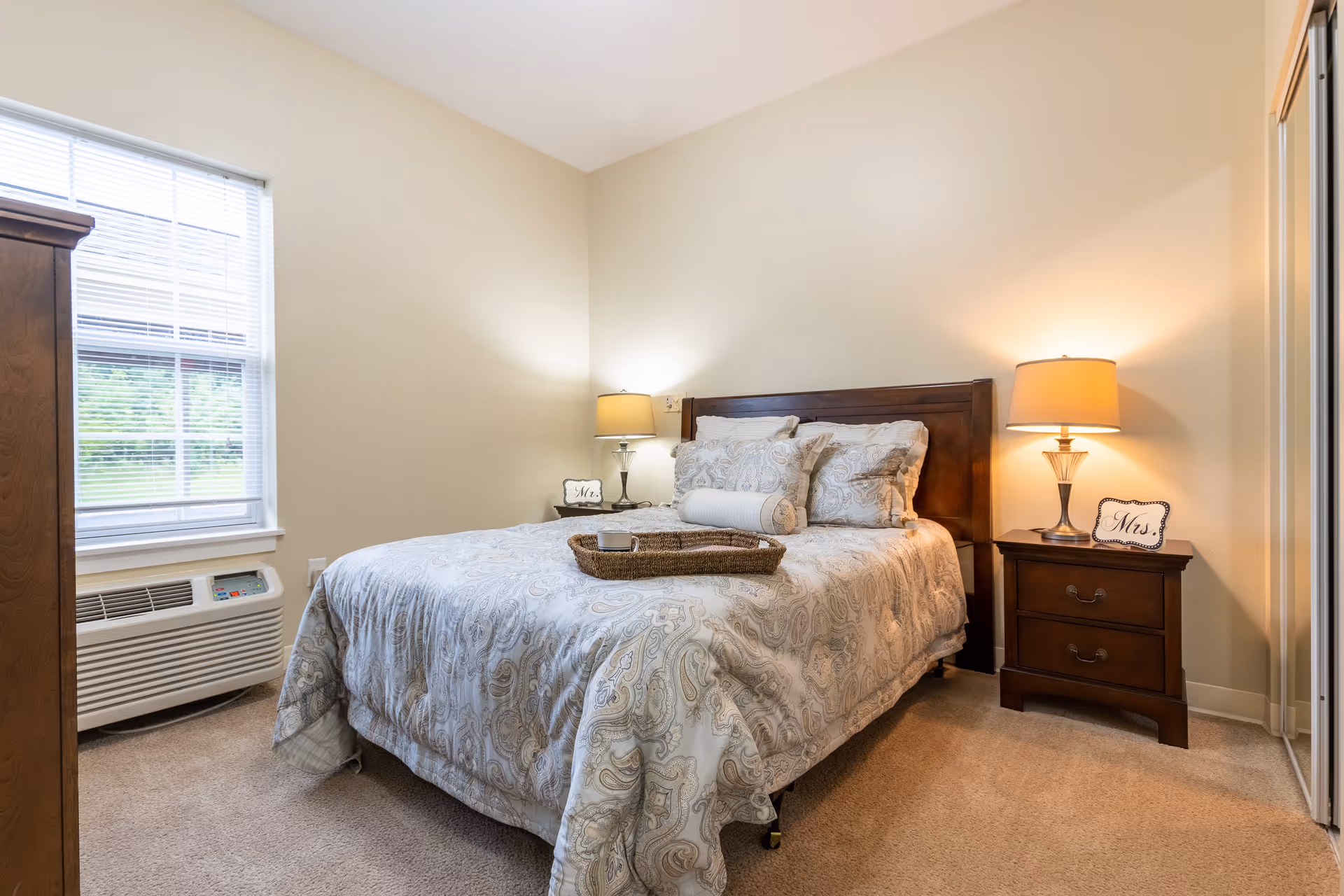 A cozy bedroom with a large bed covered in a patterned comforter and multiple pillows. There are two bedside tables with lamps on each side of the bed. One bedside table has a small sign that says 'Mr.' and the other has a sign that says 'Mrs.'. A window with blinds is on the left wall, and a heating/cooling unit is below the window. The room has beige walls and carpeted floor.