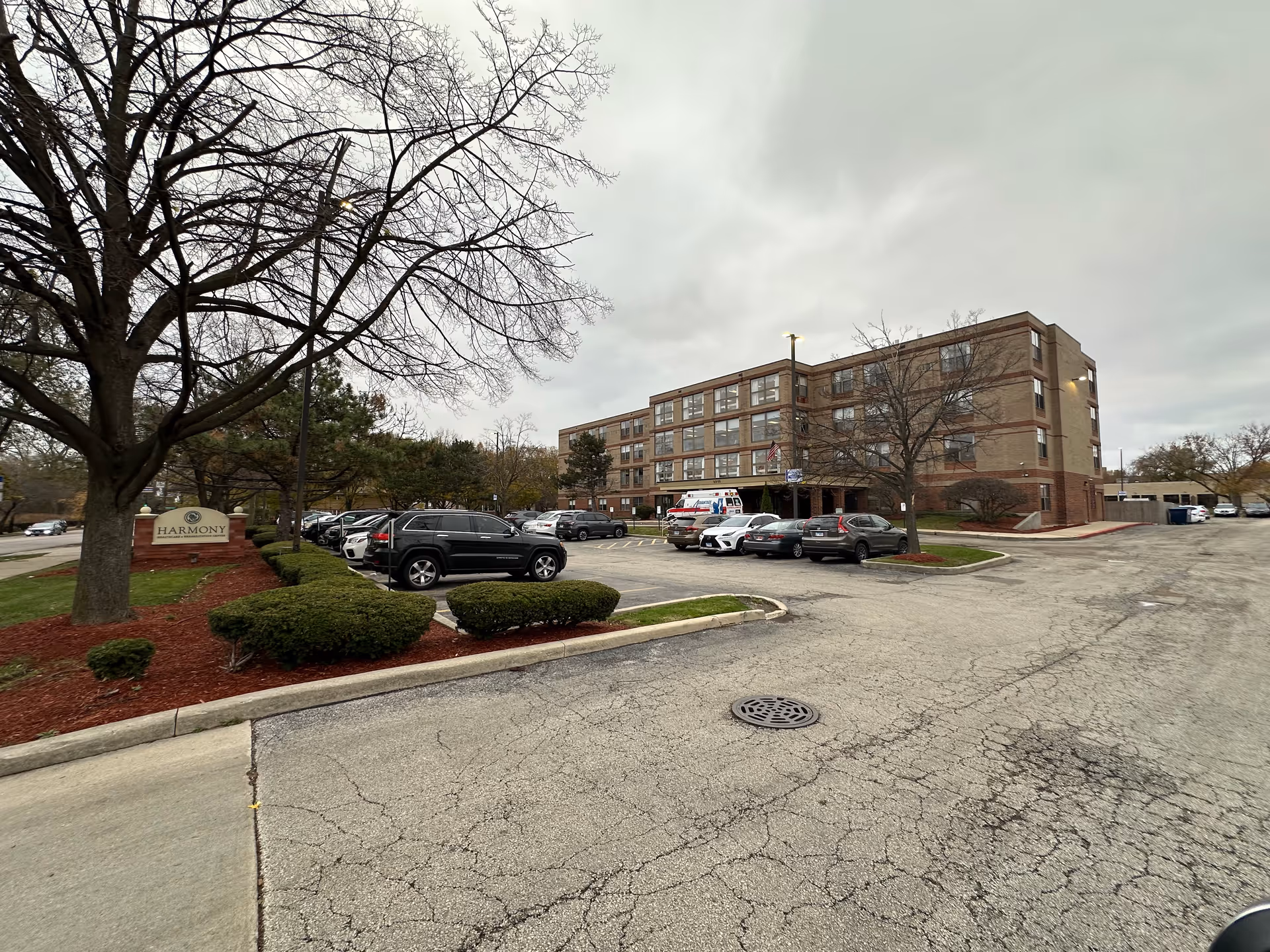 Exterior view of Harmony Healthcare & Rehabilitation Center, a multi-story brick building with numerous windows. The parking lot in front has several parked cars and an ambulance near the entrance. Leafless trees and trimmed bushes surround the area under an overcast sky.