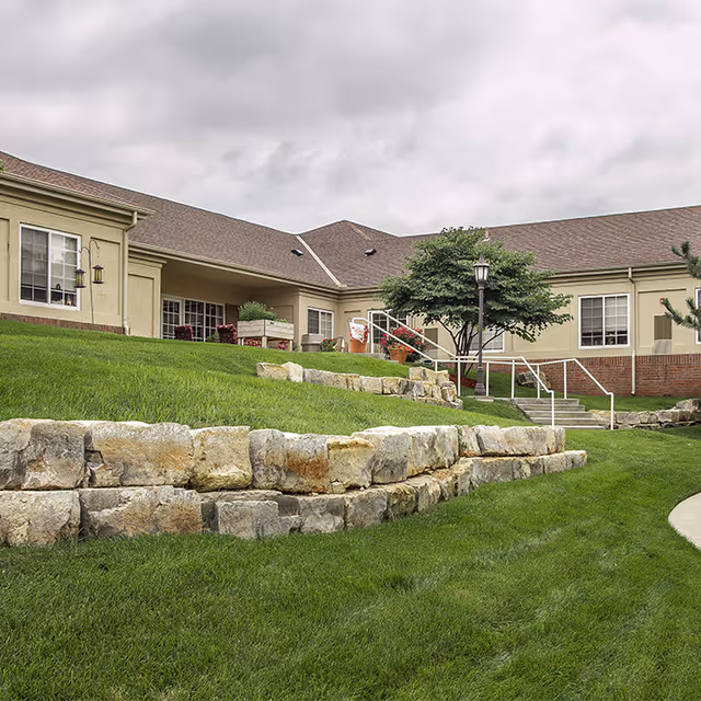 Exterior view of a senior living facility building with beige walls and brown roof, surrounded by well-maintained green grass and stone retaining walls. There are stairs with a white railing leading up to the entrance, a tree, and a lamp post near the stairs under a cloudy sky.