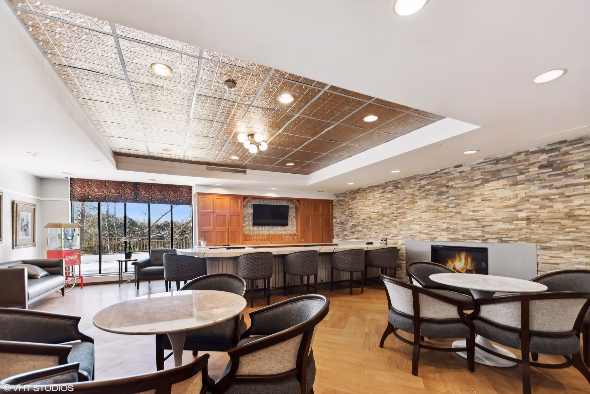 Spacious communal lounge with round marble-topped tables and chairs, a bar counter with stools, fireplace, and large windows.