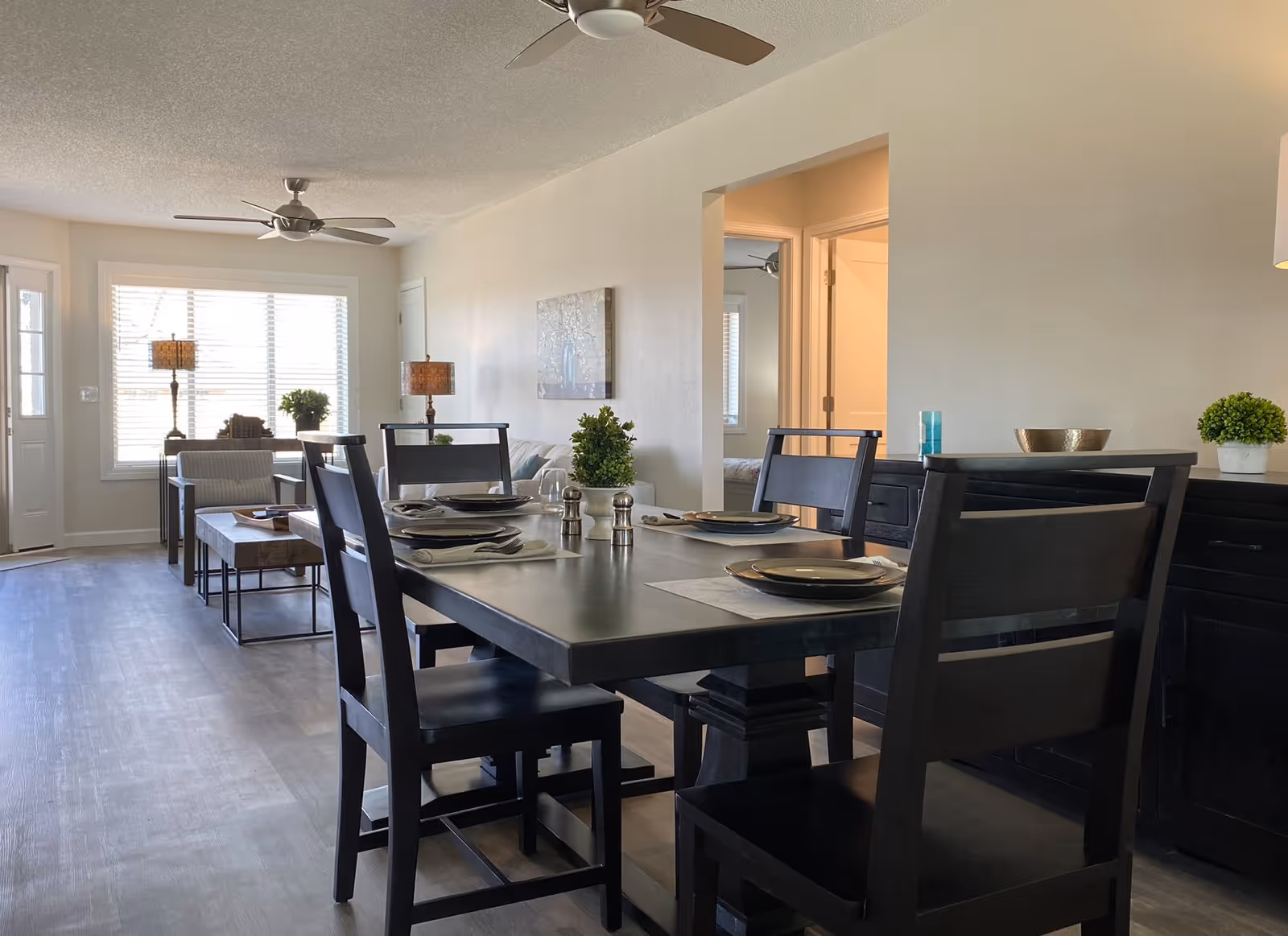 A bright and modern dining area with a dark wooden table set for four with plates and napkins. In the background, there is a living space with a sofa, two lamps on side tables, and a large window with blinds letting in natural light. The floor is a light wood laminate, and the walls are painted a soft beige. Ceiling fans are visible on the ceiling.