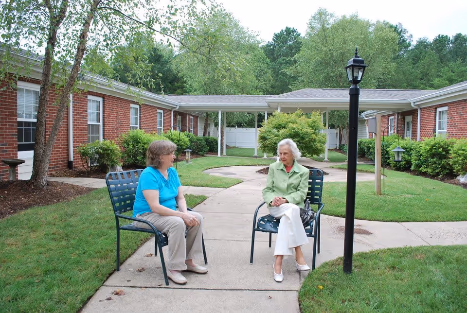 Two elderly women sitting on metal chairs in an outdoor courtyard area of a senior living facility. The courtyard has a concrete walkway, green grass, bushes, and trees. The building surrounding the courtyard is made of red brick with white window frames. A black lamp post is visible near the women.