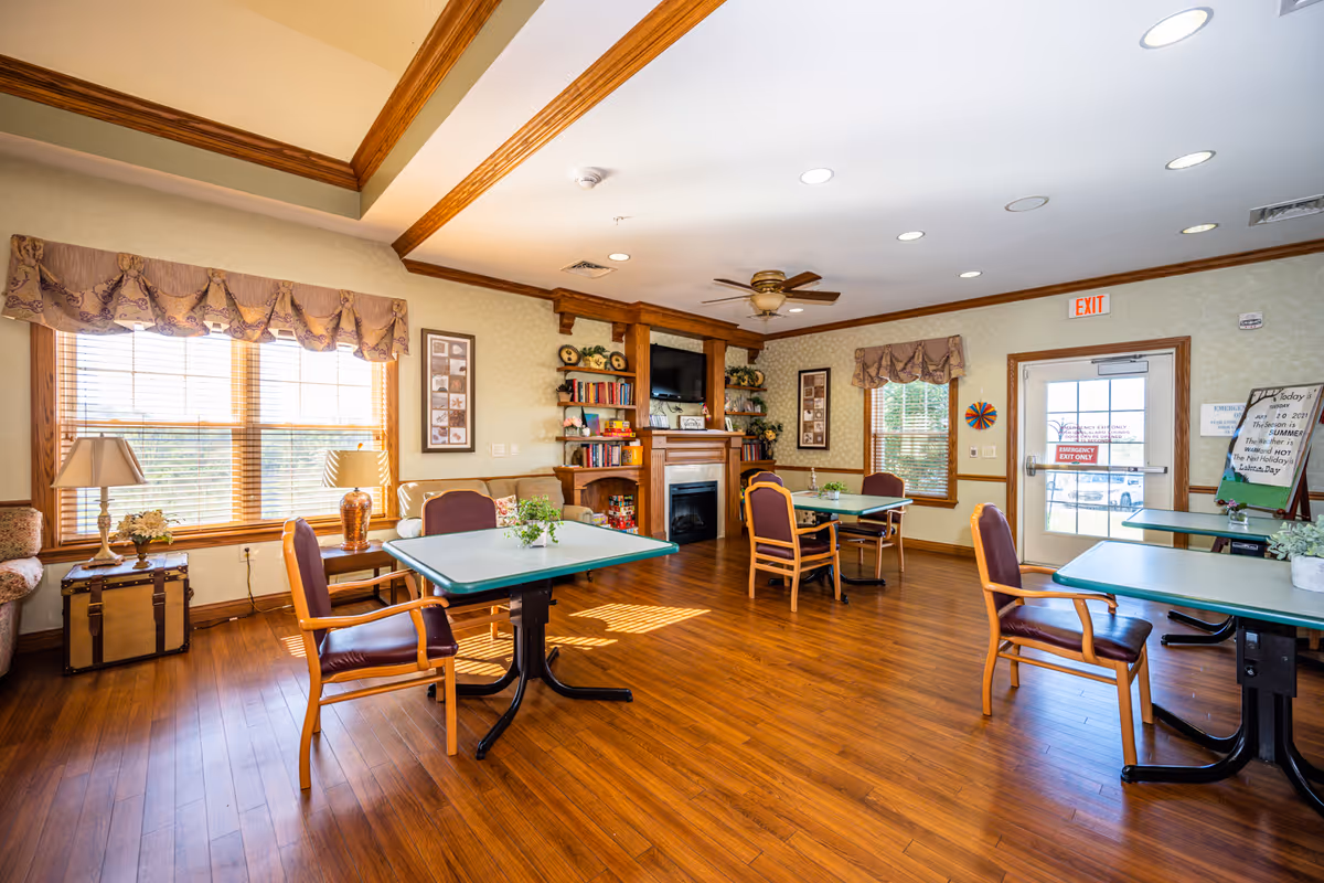 A bright and spacious common area in a skilled nursing facility featuring wooden floors, several tables with chairs, large windows with decorative valances, a fireplace with a mounted TV above it, bookshelves, and a ceiling fan. The room has warm wood trim and a door marked as an emergency exit.