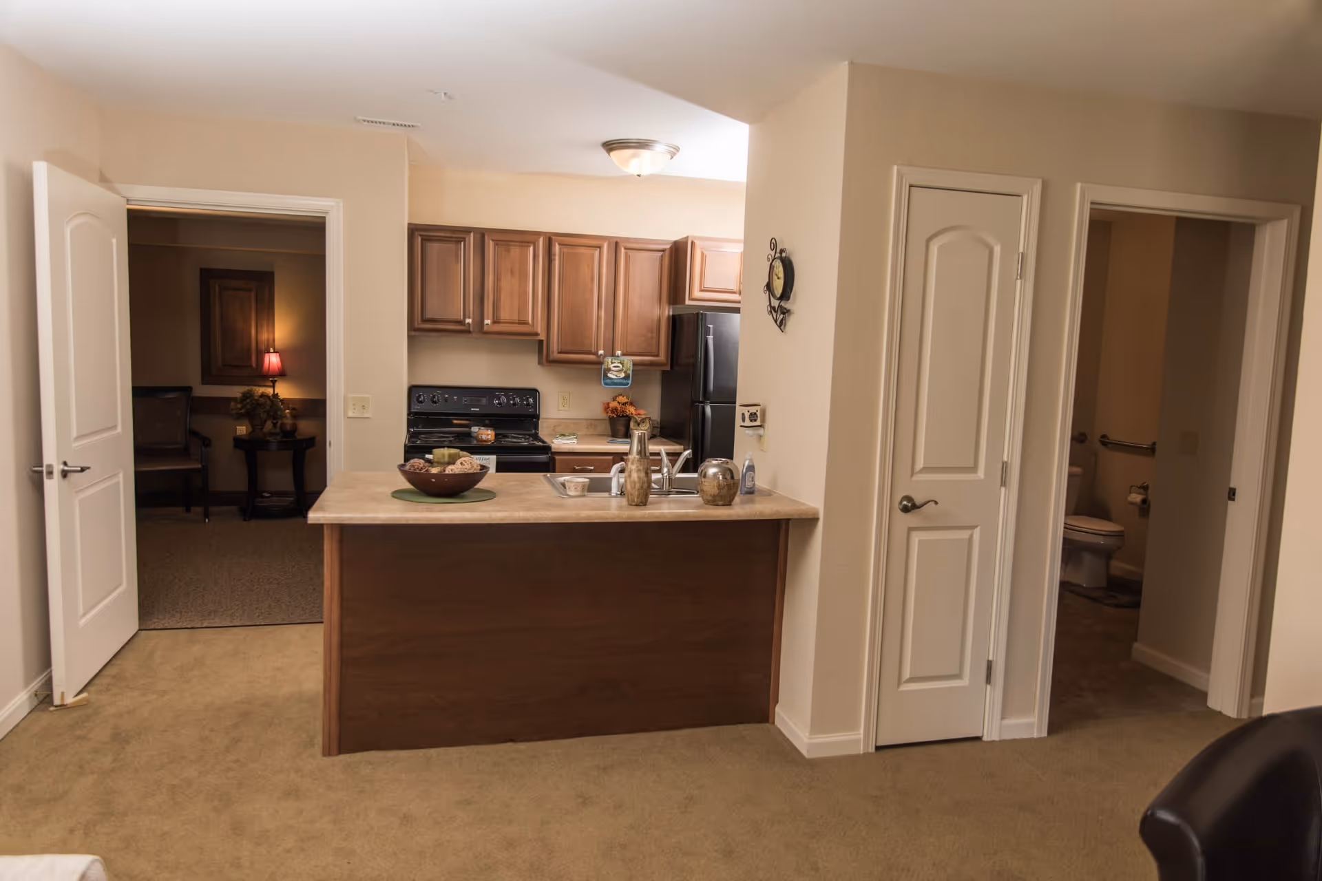 Interior view of a senior living facility apartment showing a kitchen with wooden cabinets, a black stove, and a black refrigerator. The kitchen has a countertop with decorative items. To the left, there is an open door leading to a room with a table, chair, and lamp. To the right, there are two doors, one slightly open revealing a bathroom with a toilet and grab bars. The walls are beige and the floor is carpeted.