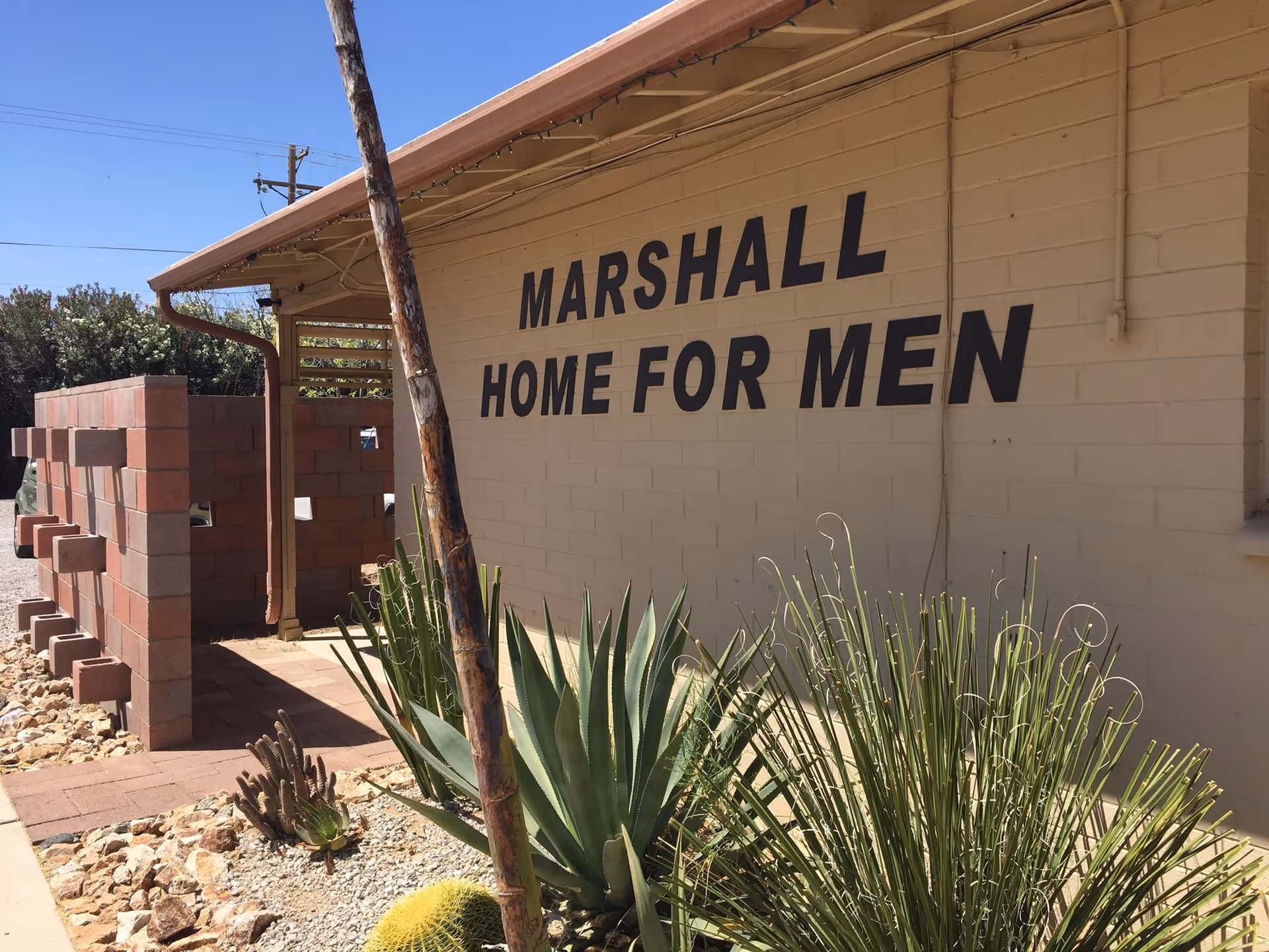 Exterior view of a single-story building with 'Marshall Home for Men' painted on the wall and desert landscaping in front.