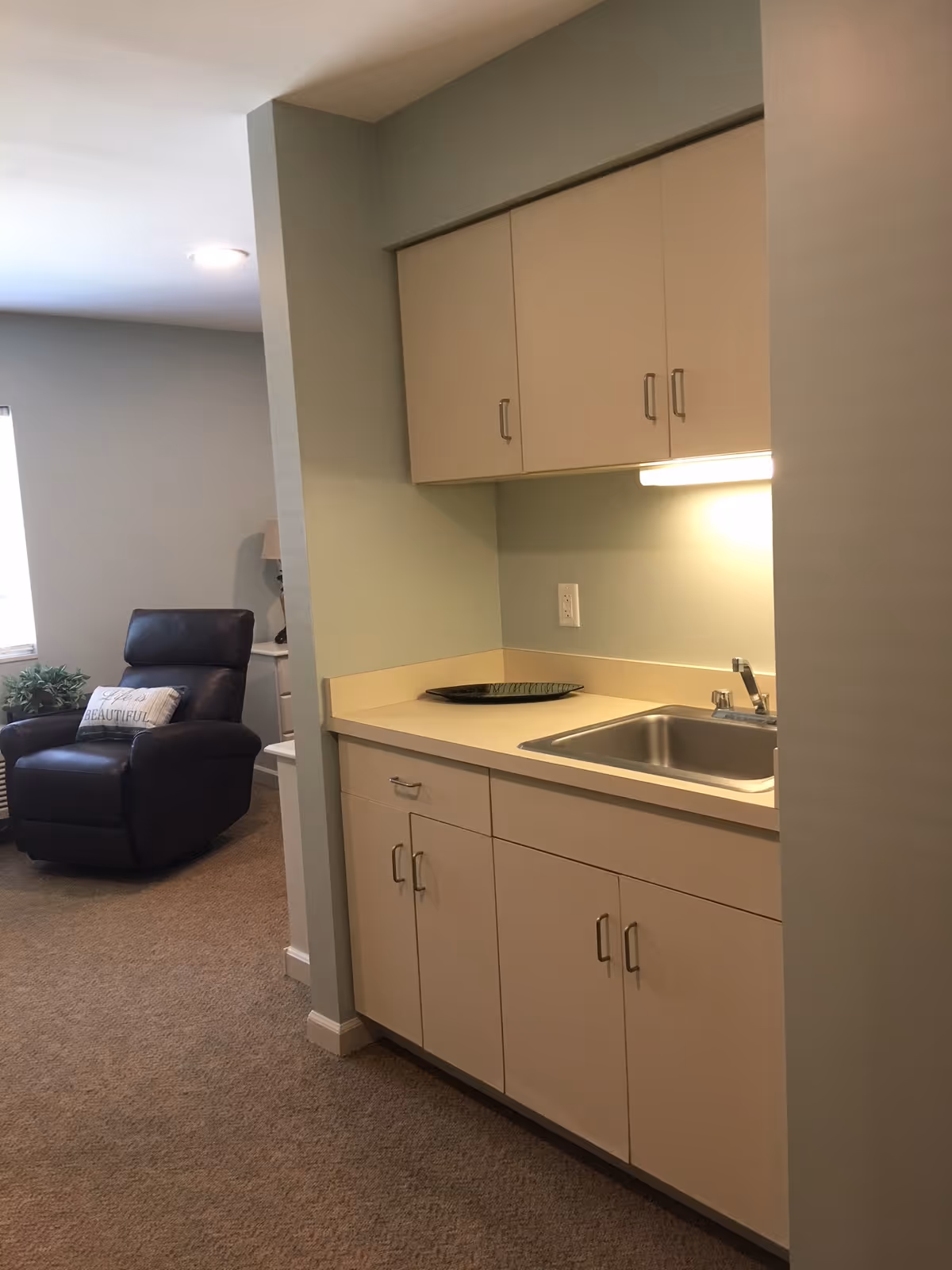 A small kitchenette area with beige cabinets, a countertop, and a stainless steel sink. There is a decorative plate on the counter. In the background, a black recliner chair with a pillow that says 'BEAUTIFUL' is visible, along with a small plant and a window letting in natural light.