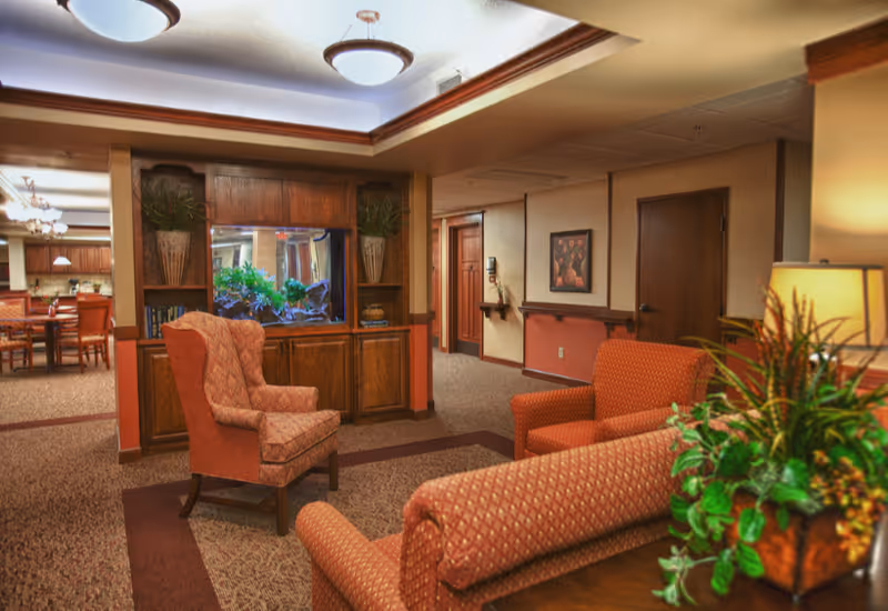 A cozy living room area in an assisted living facility featuring patterned upholstered armchairs and sofas arranged around a built-in wooden cabinet with a large aquarium. The room has warm lighting, carpeted floors, and decorative plants. In the background, a dining area with wooden tables and chairs is visible.