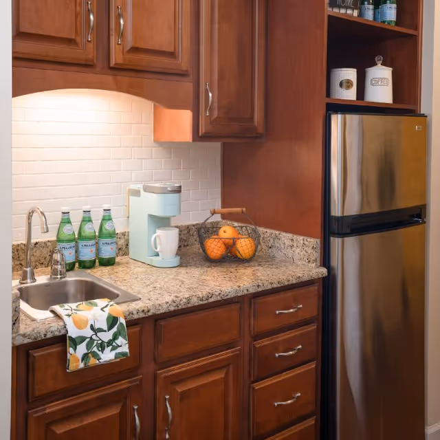 A kitchen countertop with a small sink, a coffee maker with a white mug, a basket containing oranges, and four bottles of sparkling water. The countertop is granite with wooden cabinets above and below. A stainless steel refrigerator is on the right side, and a towel with a lemon pattern hangs over the sink.